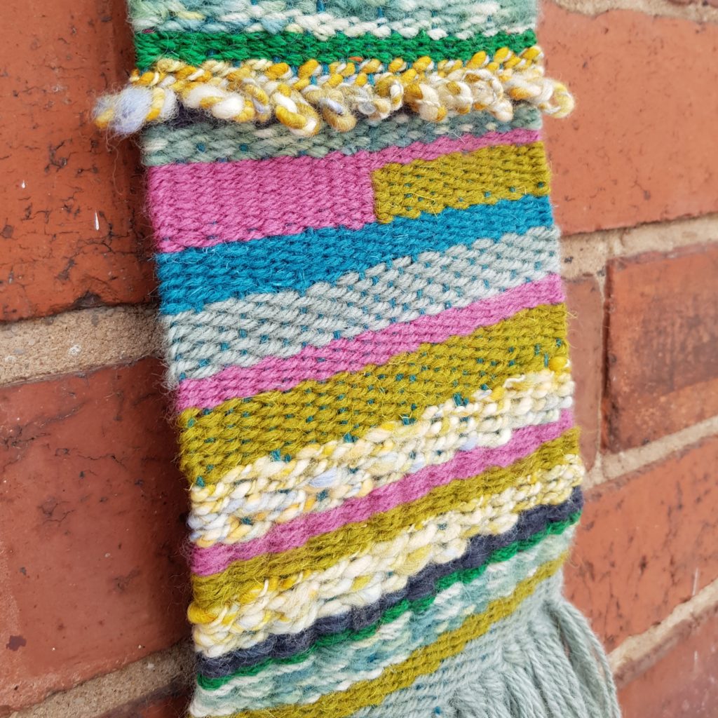 Woven Wall Hanging hung on a red brick wall. The weaving includes green, light blue, turquoise, pink and cream yarn.